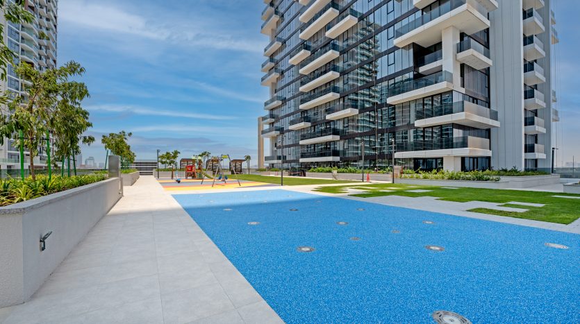 Immeuble d&#039;appartements moderne à Dubaï avec une aire de jeux colorée sur une surface en caoutchouc bleu, entourée de pelouses paysagées et de sentiers sous un ciel clair.