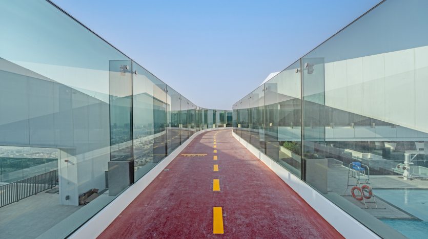 Un pont piétonnier moderne avec un sol rouge et des lignes pointillées jaunes, entouré de barrières de verre transparentes, s&#039;étendant vers un ciel bleu à Dubaï.