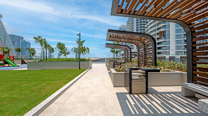 Un parc urbain moderne avec des pergolas projetant des ombres géométriques sur une allée pavée, entouré d&#039;une verdure luxuriante et d&#039;immeubles de grande hauteur contemporains, surplombant un front de mer lointain à Dubaï.