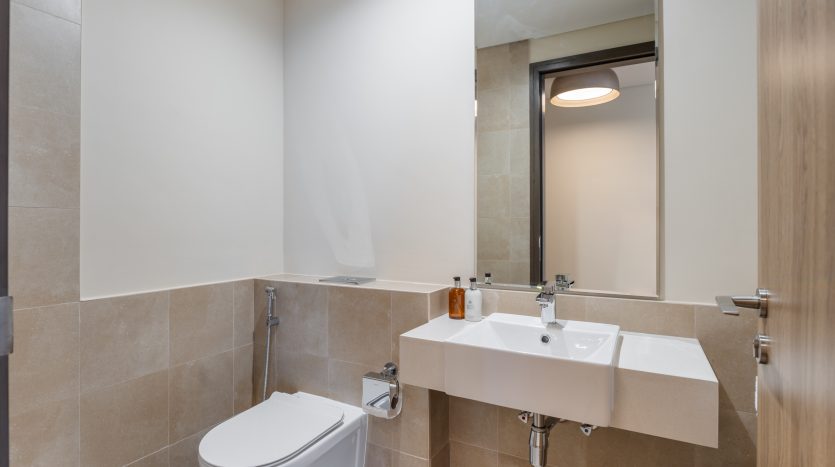 Salle de bains moderne avec carrelage beige, toilettes blanches et lavabo mural surmonté d&#039;un miroir. Une porte avec un panneau de verre réfléchissant est visible à l&#039;arrière-plan, reflétant les normes luxueuses typiques d&#039;un