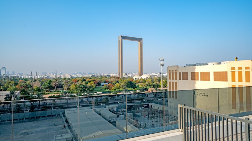 Vue depuis un balcon donnant sur un paysage urbain avec une grande structure de bâtiment unique de forme carrée au centre, entourée de bâtiments urbains et d&#039;arbres sous un ciel bleu clair à Dubaï.