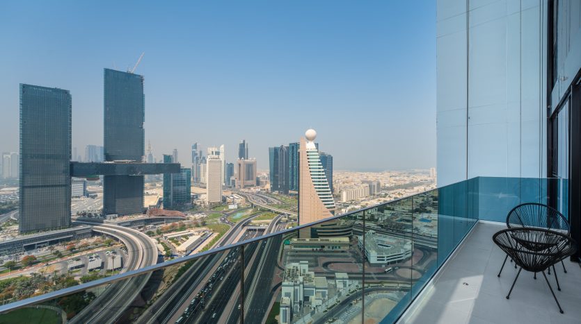 Une vue panoramique depuis un balcon de grande hauteur à Dubaï surplombant un paysage urbain moderne, avec des gratte-ciel distinctifs et un ciel bleu clair. Une seule chaise se trouve sur le balcon.