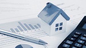 Une petite maison modèle bleue placée sur des documents financiers avec des graphiques et du texte, à côté d'une calculatrice et d'un stylo, symbolisant l'investissement immobilier à Dubaï et l'analyse financière.