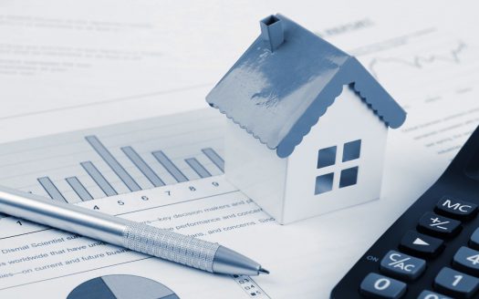 Une petite maison modèle bleue placée sur des documents financiers avec des graphiques et du texte, à côté d'une calculatrice et d'un stylo, symbolisant l'investissement immobilier à Dubaï et l'analyse financière.