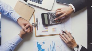 Vue de dessus d'une réunion d'affaires avec deux personnes analysant des graphiques et des données financières avec une calculatrice, un ordinateur portable et des documents sur un bureau dans une agence immobilière de Dubaï.