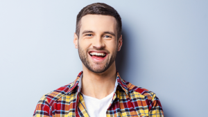 Un jeune homme joyeux avec une barbe souriant largement, portant une chemise à carreaux sur un t-shirt blanc, debout sur un fond bleu clair dans une villa de Dubaï.