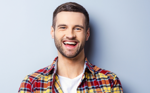 Un jeune homme joyeux avec une barbe souriant largement, portant une chemise à carreaux sur un t-shirt blanc, debout sur un fond bleu clair dans une villa de Dubaï.