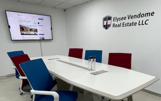 Une salle de conférence moderne comprenant une grande table blanche entourée de chaises bleues et rouges, avec un moniteur affichant au mur un site Internet de l'immobilier Dubaï marqué "elysee vendome real Estate llc.