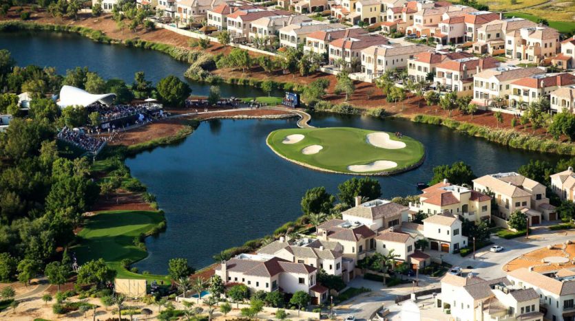 Vue aérienne d&#039;un parcours de golf verdoyant entouré d&#039;un plan d&#039;eau, avec des villas résidentielles serrées le long des bords à Dubaï, et une foule rassemblée près d&#039;un pavillon.