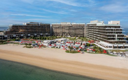 Vue aérienne d&#039;un complexe hôtelier moderne à plusieurs niveaux en bord de mer avec parasols rouges et chaises longues sur une plage de sable adjacente aux eaux bleues calmes, idéale pour un investissement à Dubaï.