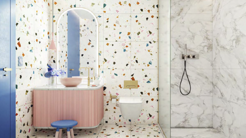 Une salle de bains lumineuse et moderne avec des murs carrelés de style terrazzo, un sol en marbre et une vasque rose avec un miroir rond. Comprend des toilettes blanches, une douche en verre et des accents décoratifs comme un tabouret bleu et
