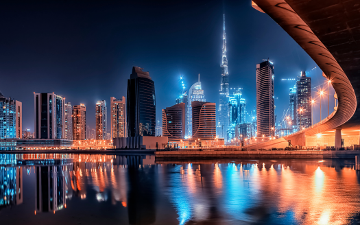 Un paysage urbain futuriste la nuit avec des gratte-ciel illuminés, une tour proéminente et un pont incurvé se reflétant dans un plan d'eau tranquille, sous un ciel bleu profond à Dubaï.