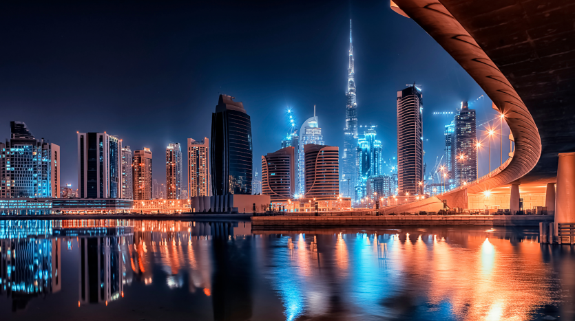 Un paysage urbain futuriste la nuit avec des gratte-ciel illuminés, une tour proéminente et un pont incurvé se reflétant dans un plan d&#039;eau tranquille, sous un ciel bleu profond à Dubaï.