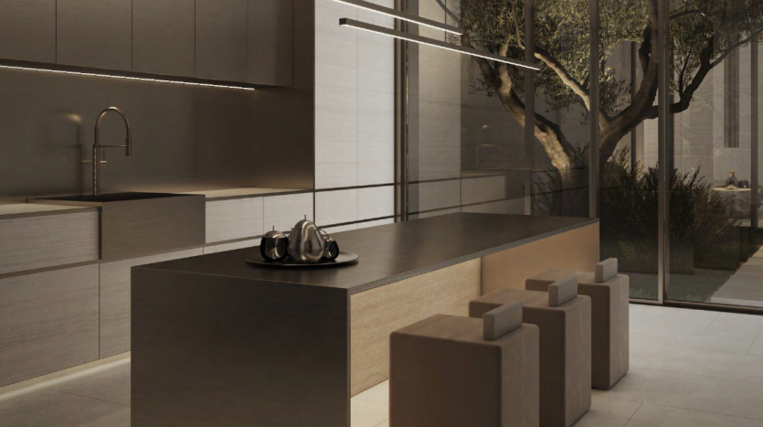 Une cuisine élégante et moderne aux finitions métalliques dans une villa de Dubaï, comprenant un îlot central, des tabourets minimalistes et un arbre intérieur visible à travers les parois vitrées.
