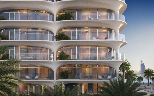 Un bâtiment moderne de plusieurs étages doté de balcons incurvés surplombe un cadre tropical avec des palmiers à Dubaï. L'extérieur est éclairé au crépuscule, mettant en valeur une ligne d'horizon urbaine en arrière-plan.