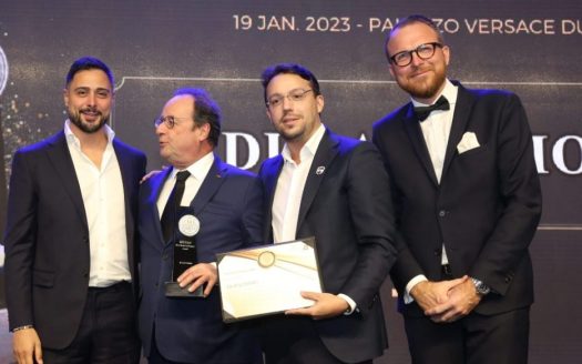 Quatre hommes en tenue de soirée, l'un titulaire d'un certificat et l'autre d'une médaille, souriant lors d'une cérémonie de remise de prix liée à l'investissement Dubaï. Détails de l'événement et date visibles en arrière-plan.