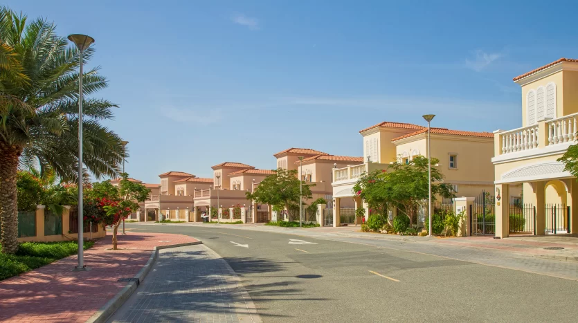 Une rue de banlieue ensoleillée de Dubaï bordée de palmiers, montrant une rangée de maisons à deux étages au toit en terre cuite avec des jardins paysagers, sous un ciel bleu clair.