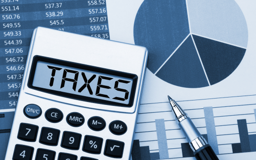 Calculatrice affichant les « taxes » sur son écran, avec un stylo, des graphiques à barres, un diagramme circulaire et des fiches de données financières sur l'investissement Dubaï en arrière-plan, représentant l'analyse financière ou le calcul des impôts.