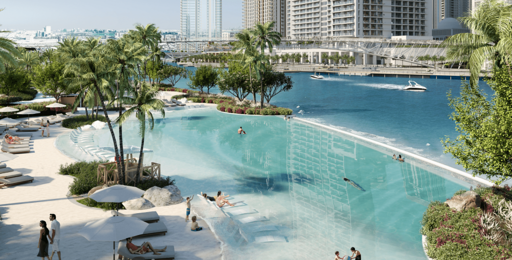 Un lujoso complejo urbano con una gran piscina azul claro, zonas de arena con tumbonas e imponentes rascacielos de fondo a orillas de un río en Dubai.