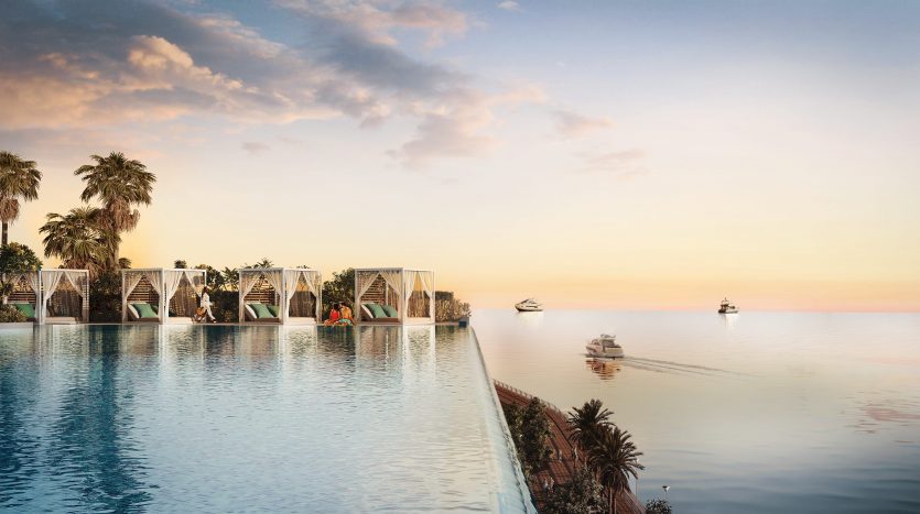 Une piscine à débordement surplombant une mer sereine au coucher du soleil, avec des palmiers et des cabines modernes semi-circulaires au bord de la piscine. Des bateaux sont visibles à l&#039;horizon sous un ciel pastel près d&#039;une luxueuse villa à