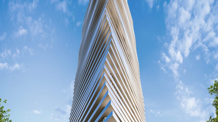 Un gratte-ciel angulaire moderne avec un design en couches, entouré d&#039;une verdure luxuriante sous un ciel bleu clair à Dubaï. La façade du bâtiment présente une série de persiennes horizontales blanches.