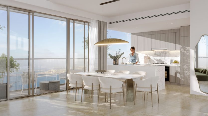 Une cuisine moderne et lumineuse avec de grandes fenêtres offrant une vue panoramique sur Dubaï. Une personne prépare à manger au comptoir. La salle à manger comprend une table ronde blanche et des chaises élégantes, avec une grande suspension.