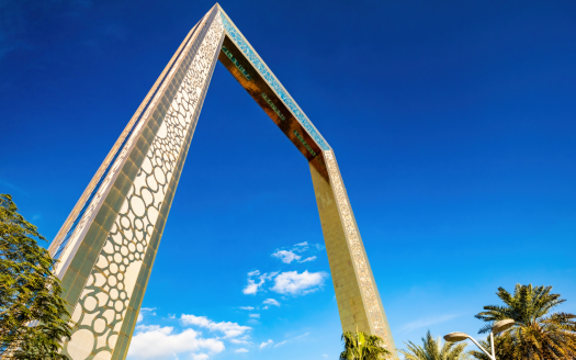 Une grande arche moderne aux motifs complexes se dresse sous un ciel bleu vif, flanquée de palmiers luxuriants près d’une villa à Dubaï. La conception de l&#039;arche comprend des détails vibrants ressemblant à des peintures murales.