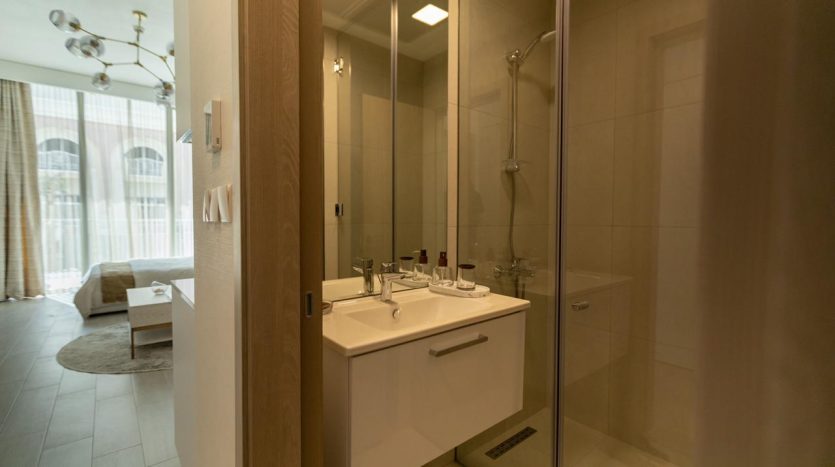 Un intérieur de salle de bains moderne comprenant une vanité blanche avec un lavabo, une cabine de douche en verre et un aperçu d&#039;un espace de vie confortable à travers la porte, illustrant une opulente propriété immobilière de Dubaï.