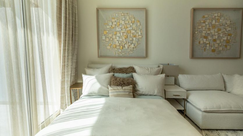 Chambre lumineuse et moderne dans une villa de Dubaï comprenant un grand lit avec une literie blanche et des oreillers moelleux, un canapé blanc et des œuvres d&#039;art murales abstraites au-dessus du lit, complétées par une douce lumière naturelle filtrant à travers le voilage.