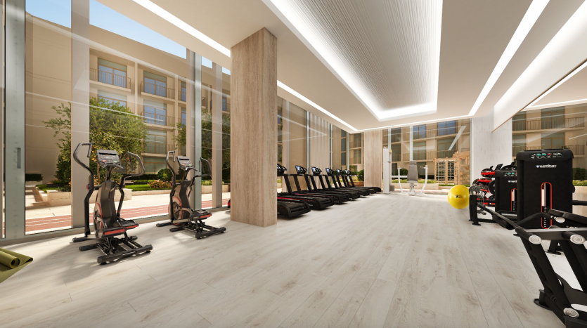 Une salle de sport spacieuse et moderne avec de grandes fenêtres, dotée de tapis roulants alignés, de machines elliptiques, de divers équipements de fitness et d&#039;une ambiance lumineuse et accueillante idéale pour investir à Dubaï.