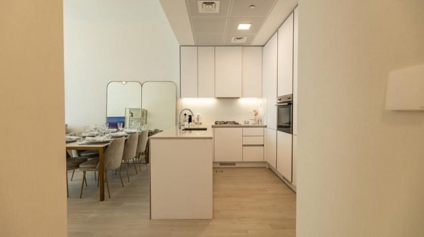 Une cuisine moderne avec des armoires blanches et des appareils électroménagers intégrés dans un appartement de Dubaï s&#039;ouvre sur un coin repas avec une table dressée, vue depuis un couloir éclairé par une lumière chaleureuse.