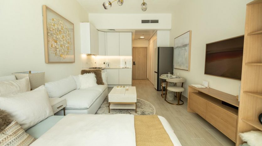 Un salon moderne dans un appartement de Dubaï avec un canapé sectionnel blanc, une console TV en bois, des œuvres d&#039;art murales abstraites, des armoires blanches et un coin repas visible en arrière-plan. La pièce adopte une couleur neutre