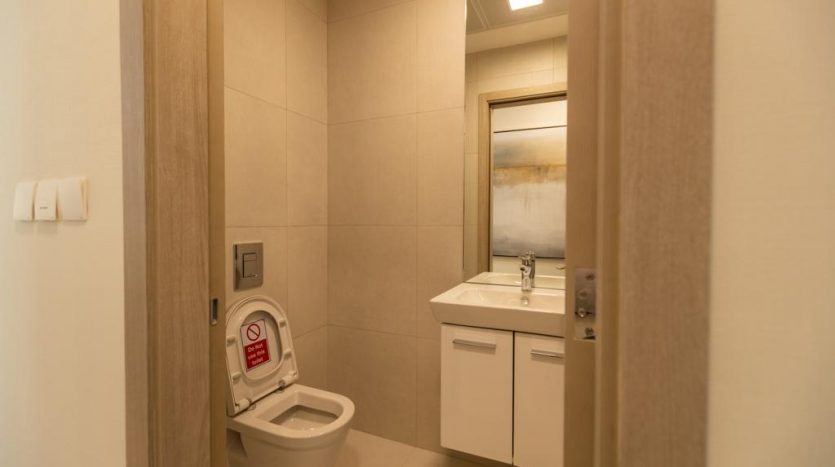 Une salle de bains moderne dans un appartement de Dubaï avec des carreaux beiges, des toilettes avec fonction bidet et un meuble lavabo blanc sous un miroir bien éclairé.