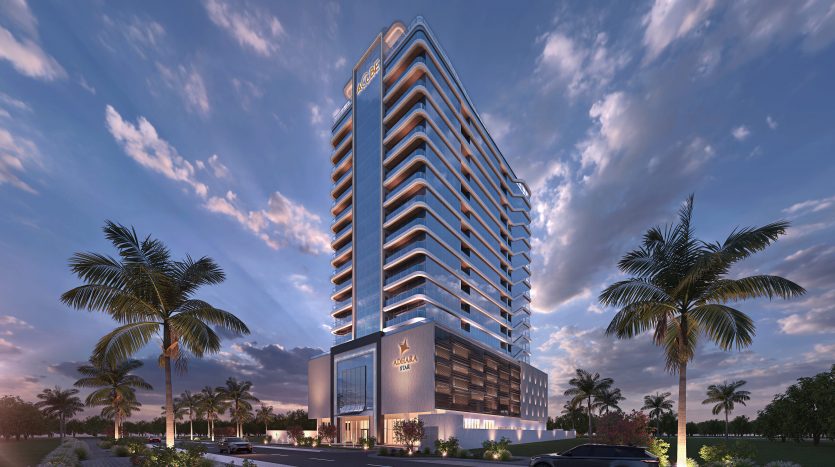 Un immeuble moderne de grande hauteur à Dubaï, illuminé au crépuscule, présentant une conception architecturale distinctive avec des balcons à chaque étage, entourés de palmiers et une entrée bien éclairée avec un logo.