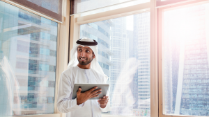 Homme émirati en tenue traditionnelle souriant et tenant une tablette, debout près d&#039;une grande fenêtre dans son appartement de Dubaï avec vue sur les gratte-ciel en arrière-plan.