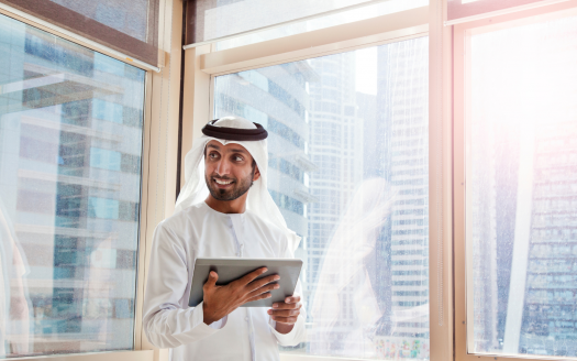 Homme émirati en tenue traditionnelle souriant et tenant une tablette, debout près d'une grande fenêtre dans son appartement de Dubaï avec vue sur les gratte-ciel en arrière-plan.