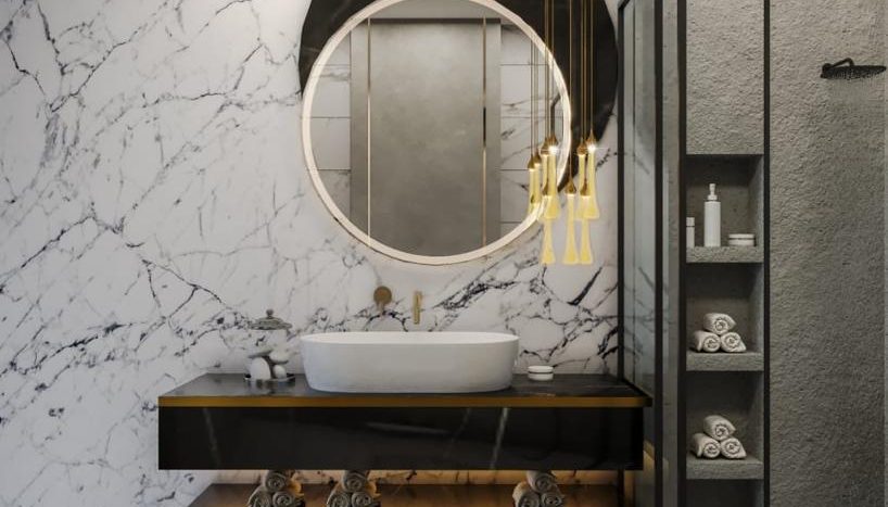 Une salle de bains moderne avec un mur et un sol en marbre, avec un miroir circulaire au-dessus d'une vanité noire contenant un lavabo blanc. Des luminaires dorés et une porte de douche en verre avec serviettes et articles de toilette visibles sont idéaux pour