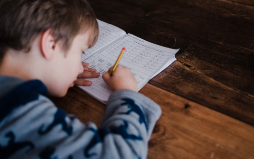 Un jeune enfant s'est concentré sur l'écriture dans un cahier avec un crayon, assis à une table en bois dans une villa à Dubaï. La vue montre le côté droit de l'enfant et le haut de sa tête.