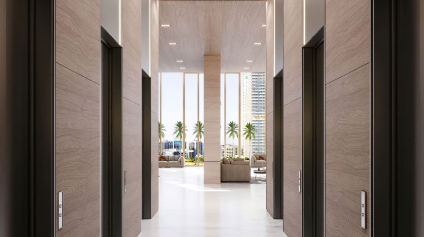 Un hall d&#039;hôtel luxueux à Dubaï avec des sols en marbre, flanqué de plusieurs ascenseurs aux murs en panneaux de bois. Le lobby se prolonge dans un espace bien éclairé avec de grandes fenêtres révélant des palmiers à l&#039;extérieur.