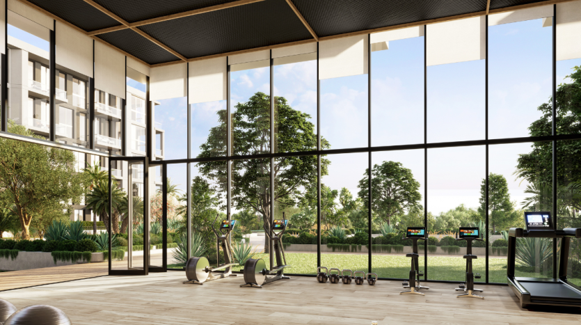 Une salle de sport moderne dotée de grandes fenêtres vitrées offrant une vue sur un jardin luxuriant et des immeubles d&#039;habitation à Dubaï. Il est équipé de tapis roulants, de poids et d&#039;un espace pour le yoga ou les étirements.