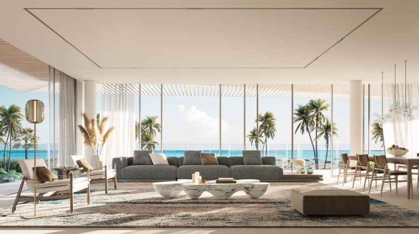 Un salon luxueux et moderne avec un grand canapé sectionnel gris, un mobilier élégant et des baies vitrées offrant une vue panoramique sur l'océan dans un appartement de Dubaï, sous un ciel lumineux et ensoleillé.