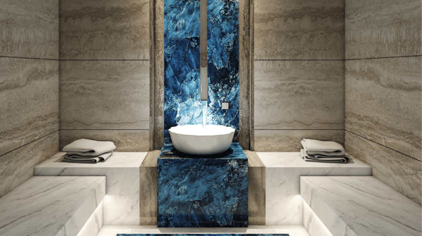 Salle de bain moderne dans une villa à Dubaï comprenant un lavabo blanc avec un robinet cascade contre un superbe mur de marbre bleu, flanqué de murs et de sols en marbre beige, avec des serviettes soigneusement pliées sur le côté.