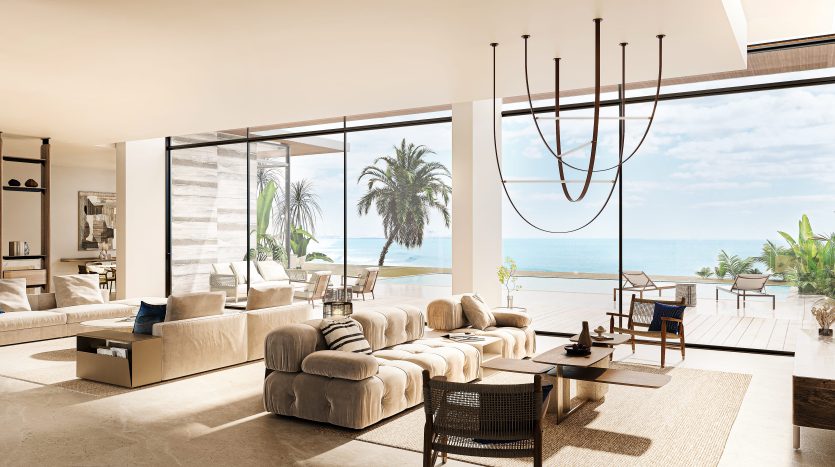 Luxueux salon décloisonné dans une villa de Dubaï avec de grandes fenêtres révélant une vue sereine sur la plage, un mobilier moderne et un lustre décoratif, mêlant harmonieusement confort intérieur et paysage tropical.