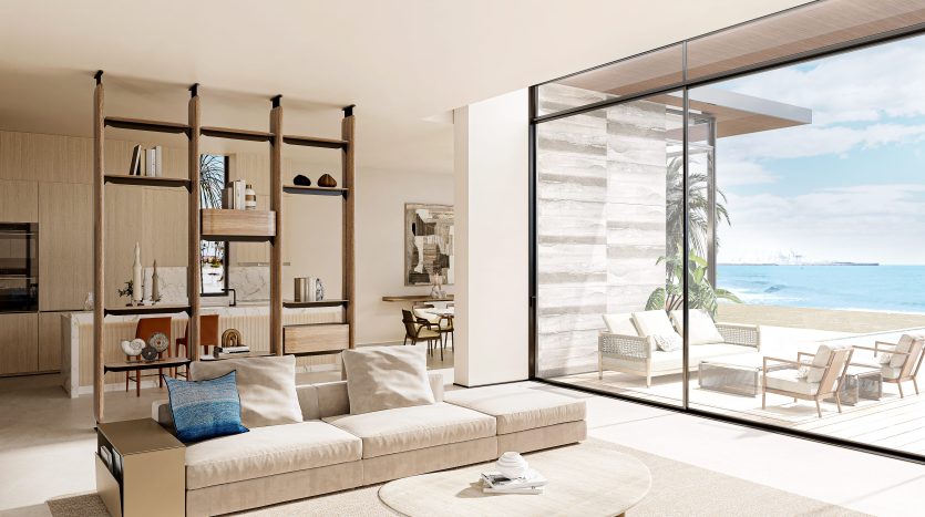 Salon moderne en bord de mer avec de grandes portes coulissantes en verre ouvrant sur un patio et une vue sur l'océan dans un appartement de Dubaï, doté d'un mobilier élégant, d'étagères en bois et d'un design minimaliste.