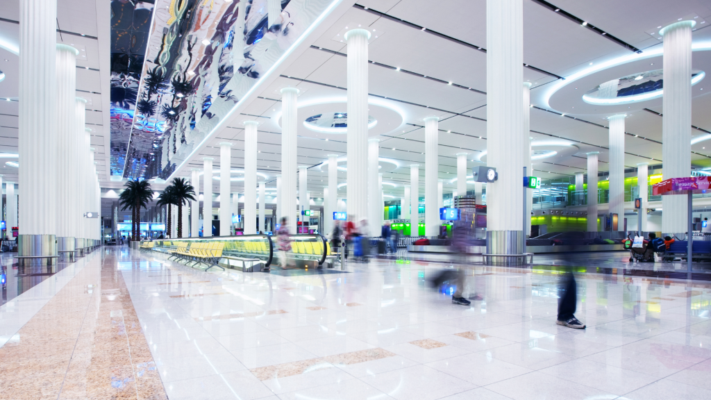 Terminal d'aéroport moderne et animé avec des silhouettes floues marchant, des colonnes blanches, un éclairage vif et des sols brillants réfléchissants gérés par l'agence immobilière de Dubaï.