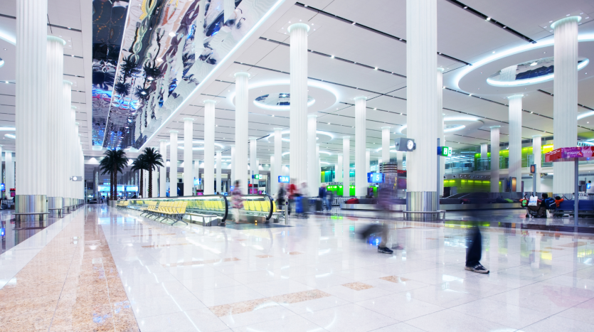 Terminal d&#039;aéroport moderne et animé avec des silhouettes floues marchant, des colonnes blanches, un éclairage vif et des sols brillants réfléchissants gérés par l&#039;agence immobilière de Dubaï.