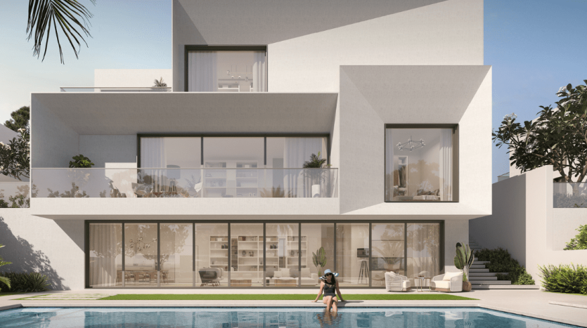 Une villa blanche moderne à plusieurs niveaux avec de grandes fenêtres, des balcons et une piscine. Une personne se promène au bord de la piscine entourée d’une verdure luxuriante. Cette propriété est répertoriée par un top AG