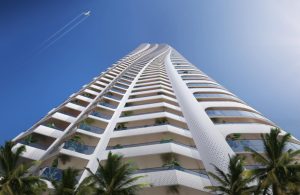 Un immeuble moderne de grande hauteur avec de vastes balcons incurvés sous un ciel bleu, flanqué de palmiers à Dubaï. Un jet laisse une trace dans le ciel clair au-dessus.