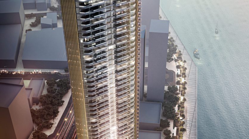 Un gratte-ciel rendu numériquement situé au bord de l&#039;eau à Dubaï, présentant un extérieur élégant et doré avec un design ondulé, à côté d&#039;un paysage urbain animé avec des bateaux dans l&#039;eau.