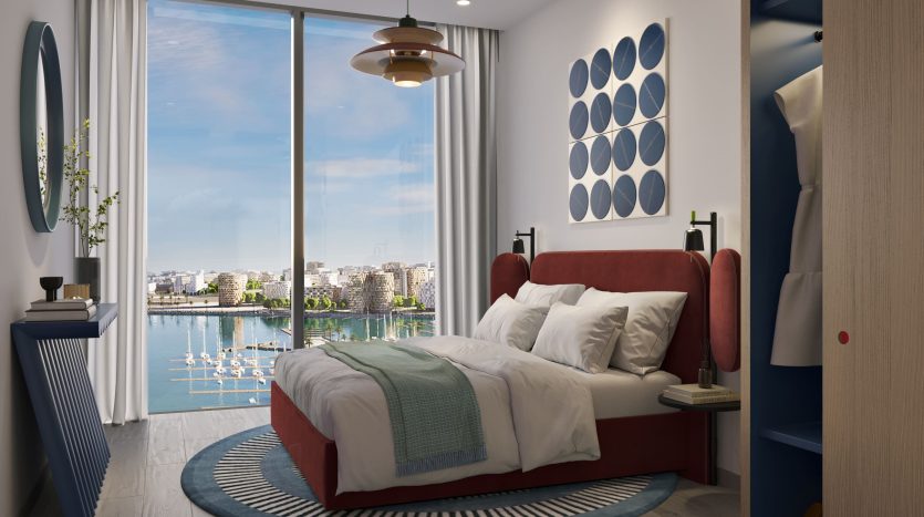 Chambre moderne dans un appartement de Dubaï avec une grande fenêtre donnant sur un paysage urbain et sur l&#039;eau. Comprend un lit rouge avec une literie blanche, un plafonnier rond, des touches bleues et une décoration minimaliste.